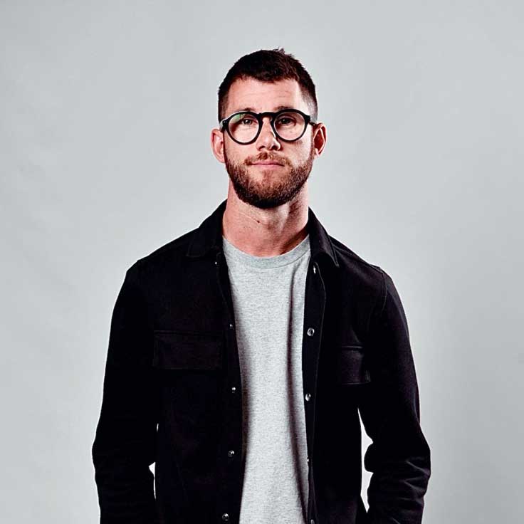 A man wearing a black shirt and a grey T-shirt stands against a neutral background.
