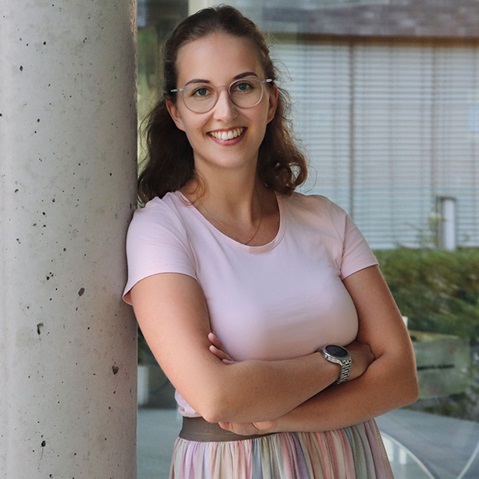 A woman leans casually against a column, wearing a pink t-shirt and a long striped skirt.