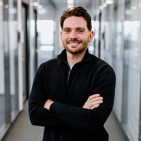 Eine Person in einem schwarzen Reißverschluss-Sweater steht mit verschränkten Armen in einem modernen Flur mit Glaswänden.