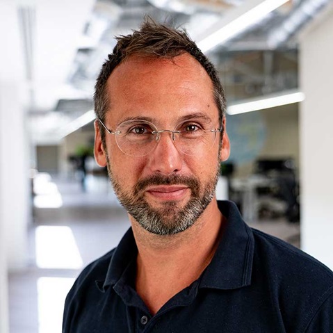 Ein Mann mit kurzem braunem Haar und leichtem Bart trägt ein dunkles Poloshirt in einem hellen Großraumbüro mit Schreibtischen, Deckenleuchten und sichtbaren Brillenbügeln.