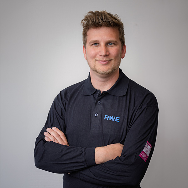 Eine Person in einem schwarzen Poloshirt mit einem blauen RWE-Logo, die mit verschränkten Armen vor einem hellen Hintergrund steht. Portrait von Daniel Helmig.