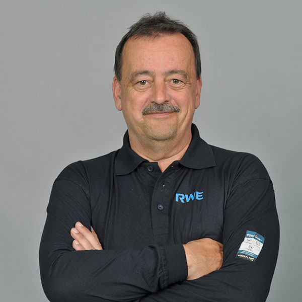 Eine Person in einem schwarzen Poloshirt mit RWE-Logo, die mit verschränkten Armen vor einem grauen Hintergrund steht.
