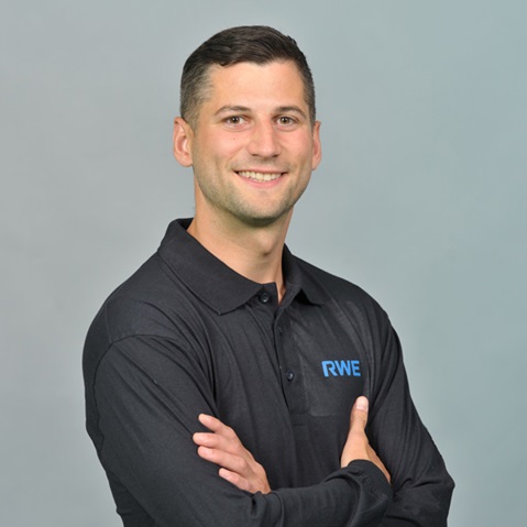Eine Person in einem schwarzen Poloshirt mit einem RWE-Logo, die mit verschränkten Armen vor einem grauen Hintergrund steht. Portrait von Dirk Decomain.