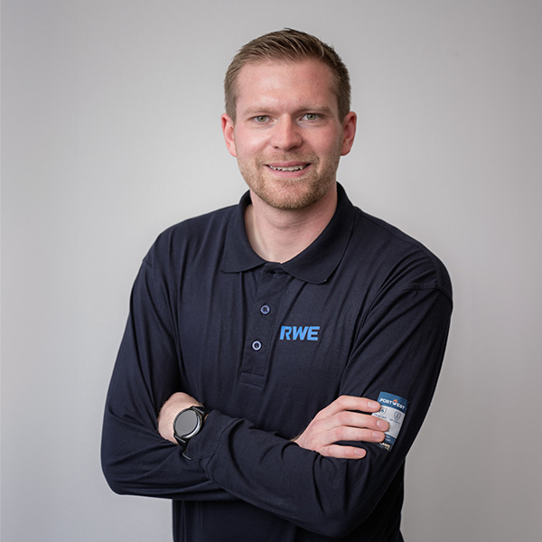Ein Mann in einem dunklen Poloshirt mit RWE-Logo steht mit verschränkten Armen vor einem neutralen Hintergrund. Portrait von Florian Hensen.