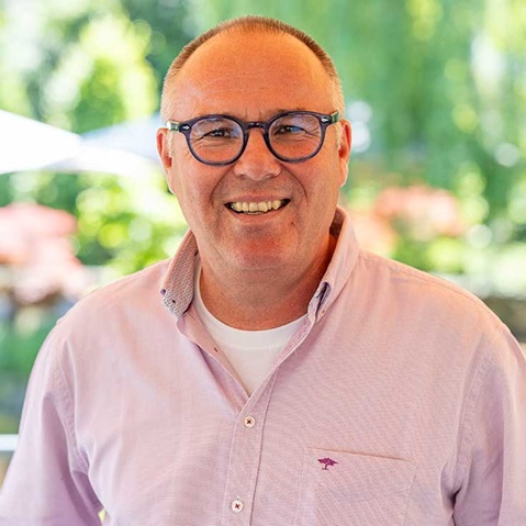 Eine Person trägt ein hellrosa Hemd und steht im Freien vor einer grünen, verschwommenen Landschaft.