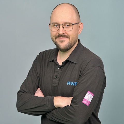 Eine Person in einem schwarzen Vans-Shirt, mit heller Haut, die vor einem grauen Hintergrund steht und Ohrringe trägt. Portrait von Jan Wilke.