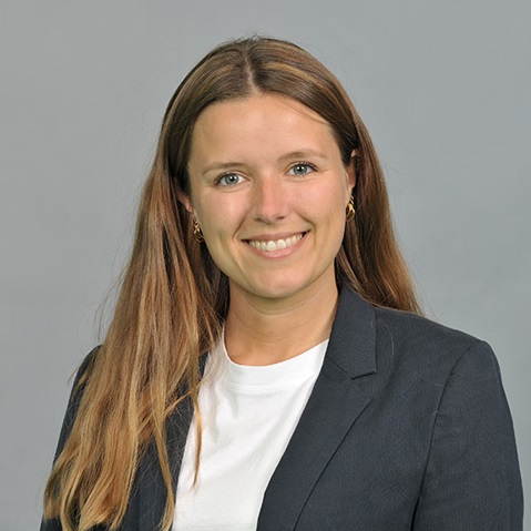 Eine Frau mit langen Haaren, die ein elegantes Jackett über einem weißen Oberteil trägt, vor einem einfarbigen grauen Hintergrund posiert.