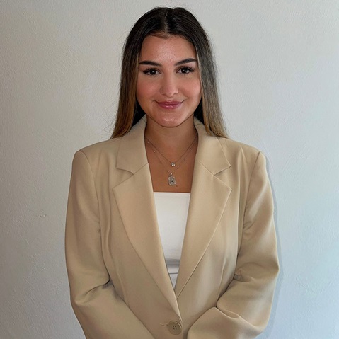 Eine Frau in einem beigen Blazer und einem weißen Oberteil steht vor einer hellen Wand und trägt Halsketten.