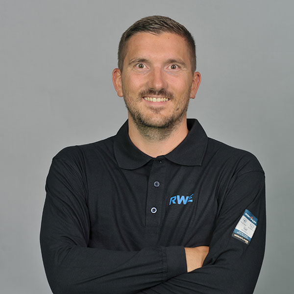 Eine Person in einem schwarzen Poloshirt mit 'RWE'-Logo steht mit verschränkten Armen. Der Hintergrund ist grau.