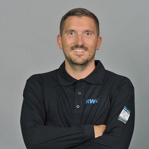 Eine Person in einem schwarzen Poloshirt mit 'RWE'-Logo steht mit verschränkten Armen. Der Hintergrund ist grau.