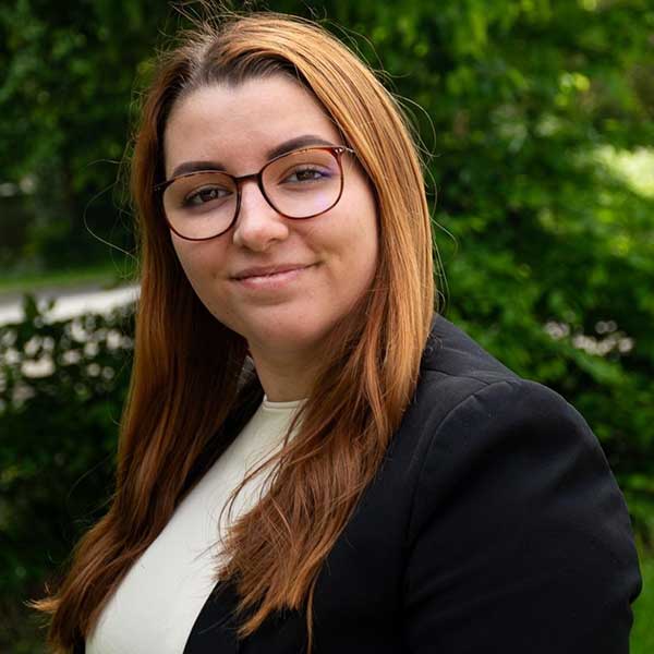 Eine Frau in einem grauen, gemusterten Blazer und einem weißen Oberteil steht vor einem neutralen Hintergrund.