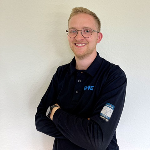 Ein lächelnder junger Mann mit kurzen blonden Haaren und runder Brille steht mit verschränkten Armen in einem dunkelblauen Poloshirt vor einer hellen Wand.
