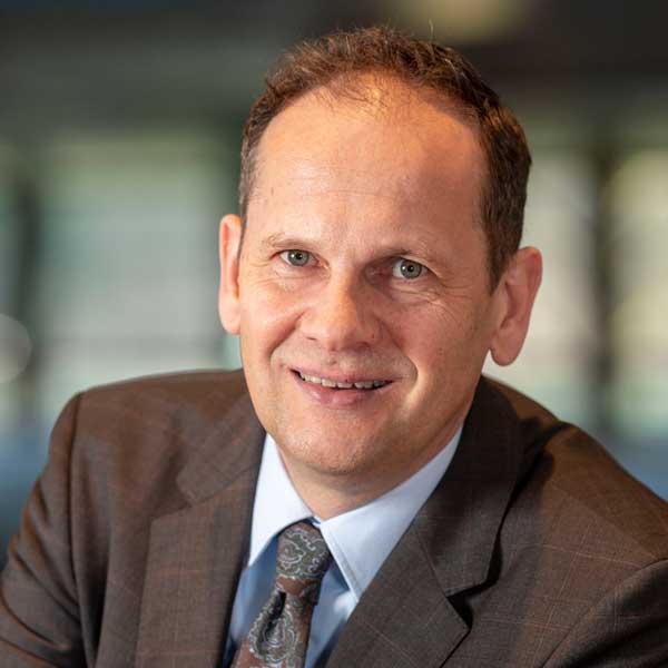 Ein Mann in einem braunen Anzug und einer gemusterten Krawatte, der an einem Schreibtisch sitzt, mit einem verschwommenen Hintergrund.