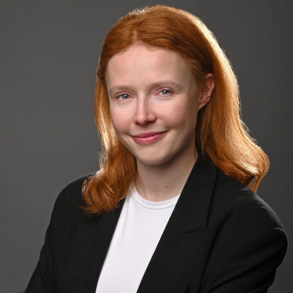 Eine Person mit langen roten Haaren trägt ein schwarzes Blazer und ein weißes Hemd, posiert vor einem dunklen Hintergrund.