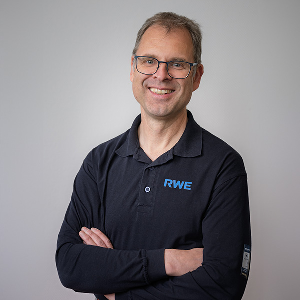 Ein Mann steht mit verschränkten Armen und trägt ein schwarzes Poloshirt mit einem blauen RWE-Logo vor einem grauen Hintergrund. Portrait von Thomas Daun.