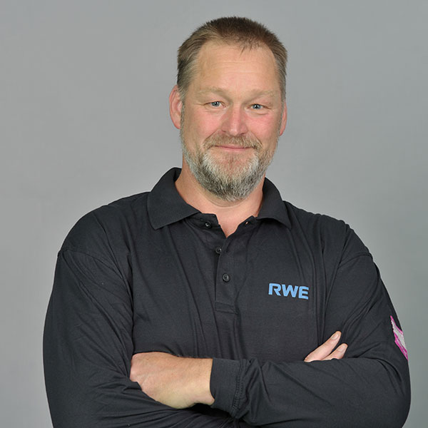 Eine Person in einem schwarzen Langarm-Polohemd mit RWE-Logo steht selbstbewusst mit verschränkten Armen vor einem grauen Hintergrund.