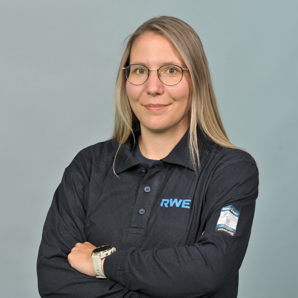 Eine Frau mit langen Haaren trägt ein schwarzes RWE-Polohemd, die Arme verschränkt, vor einem hellgrauen Hintergrund. Portrait von Vanessa Scheffler.