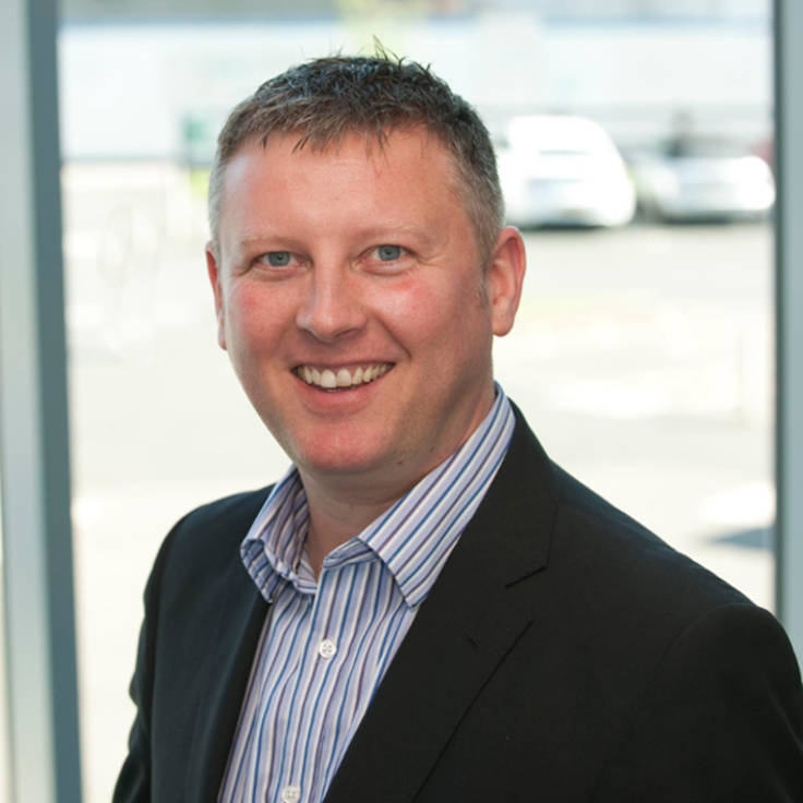 A man in a suit stands by a window with a blurred face. He is wearing a striped shirt.