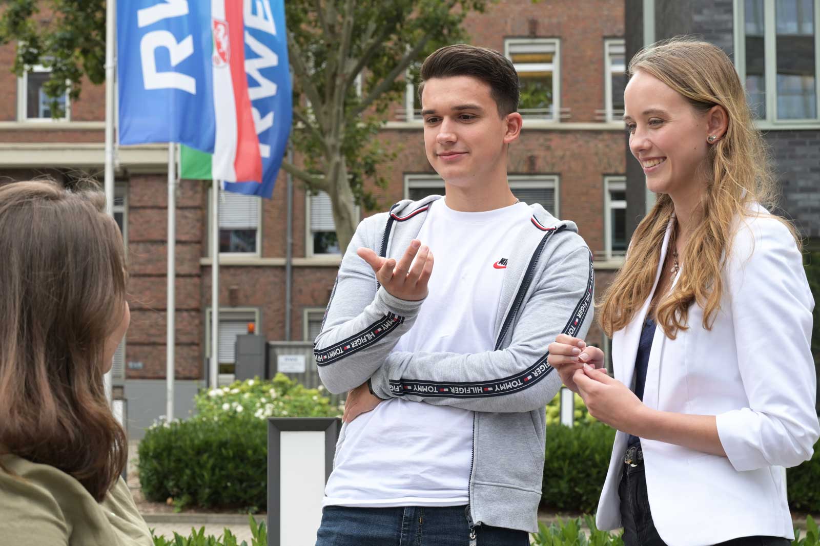 Drei junge Menschen führen ein Gespräch im Freien, mit im Hintergrund wehenden Fahnen und einem modernen Gebäude.