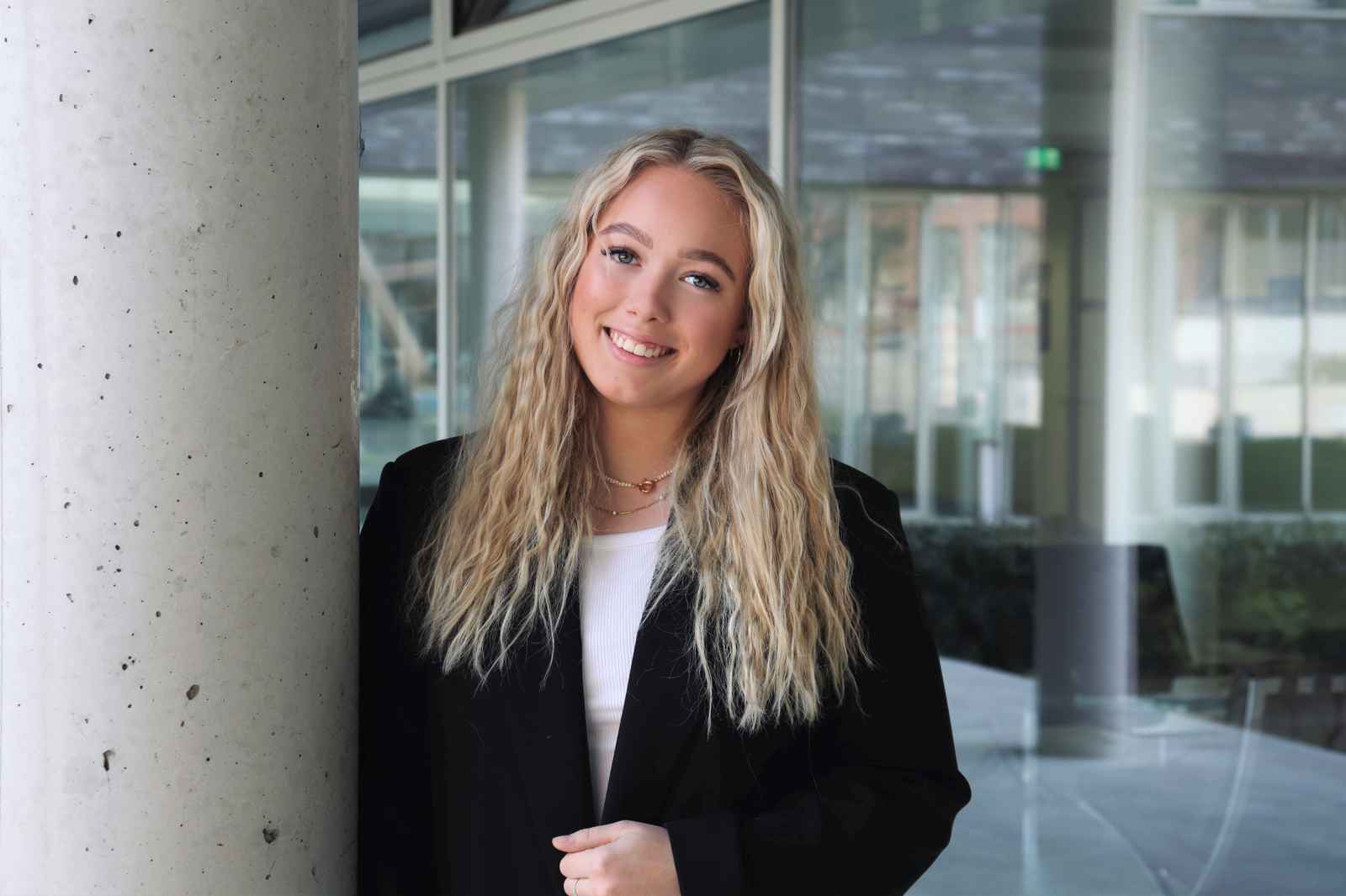Eine Person mit langen, lockigen Haaren steht neben einer Betonsäule in einem modernen Gebäude, gekleidet in einem schwarzen Mantel.