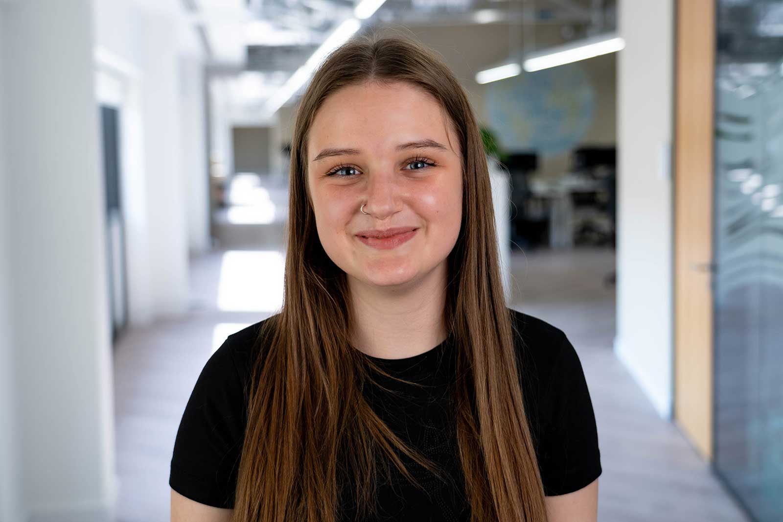 A person with long brown hair is wearing a black t-shirt, standing in a bright office space with a modern design.