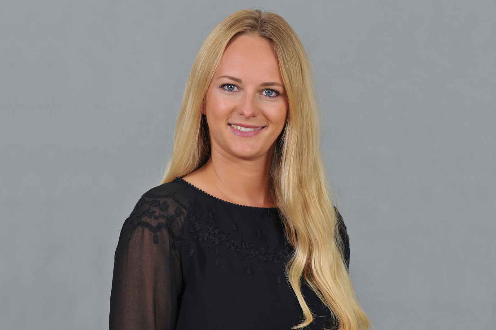 Eine Person mit langen, blonden Haaren trägt ein schwarzes Oberteil mit Spitzenakzenten vor einem grauen Hintergrund.