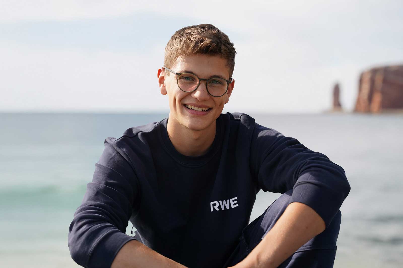 Eine Person sitzt am Meer, trägt einen navyblauen Sweatshirt mit RWE-Logo, vor einer Klippenlandschaft und bewölkten Himmel.