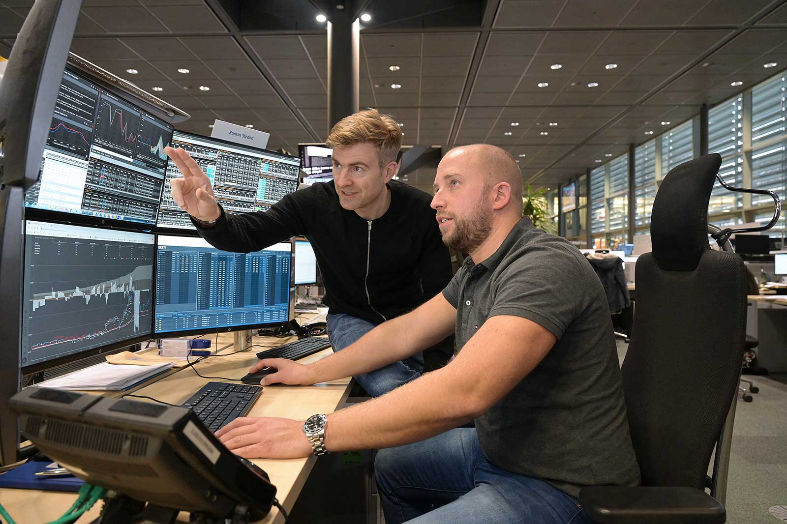 Zwei Männer im Büro analysieren Finanzdaten auf mehreren Bildschirmen. Der eine zeigt auf Grafiken, während der andere tippt.