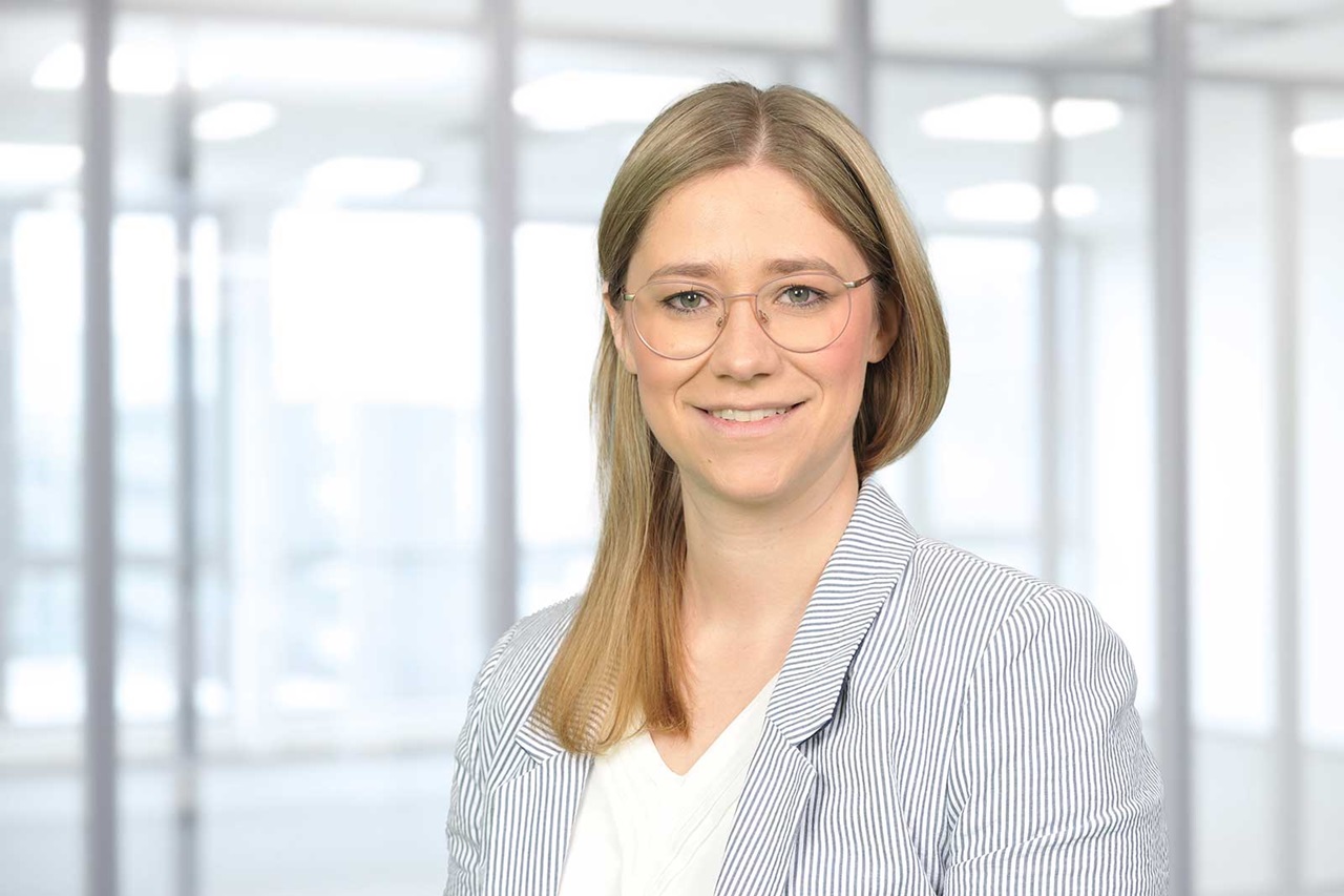 Eine Frau mit blonden Haaren trägt eine gestreifte Jacke und steht in einem hellen, modernen Büro.