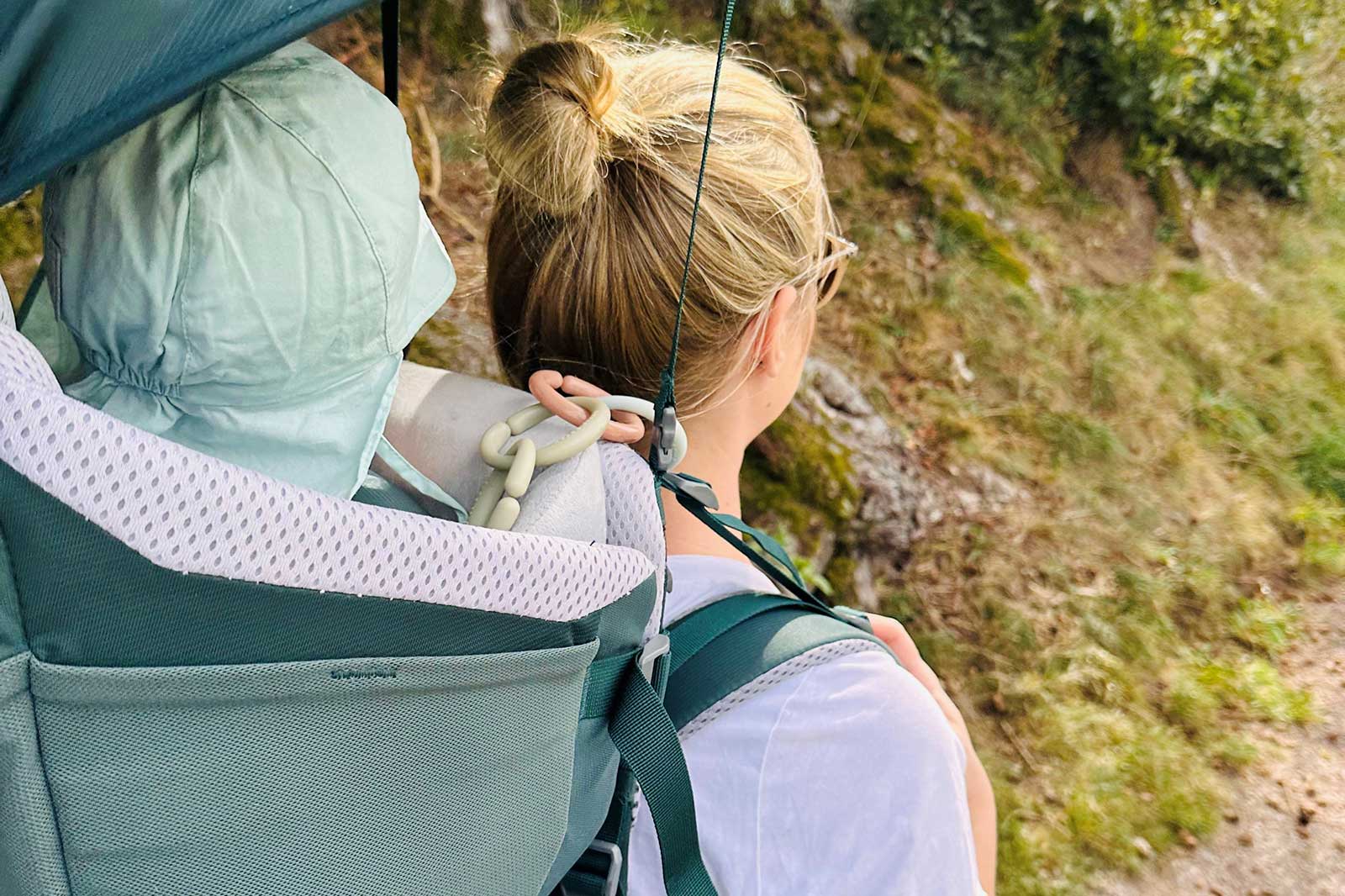 A woman carries a baby in a backpack carrier. The baby is wearing a green hood and the surroundings are natural.