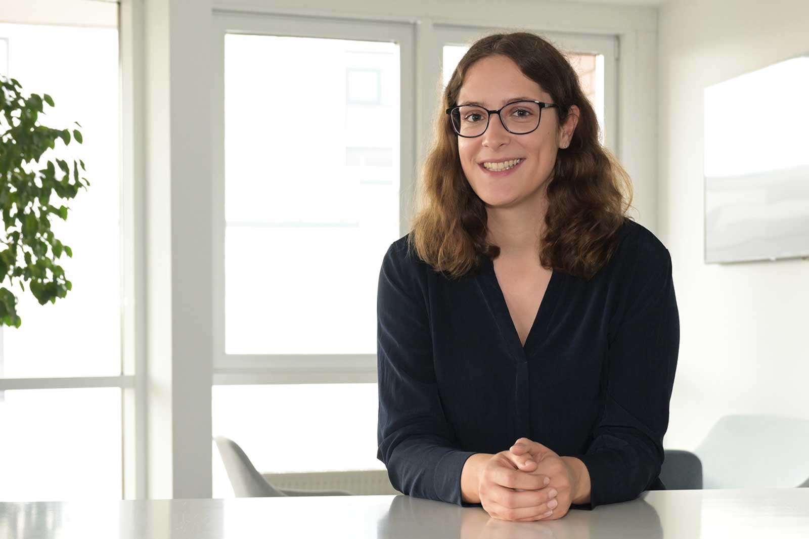 A person sits at a table in a bright room with large windows and a plant in the background.