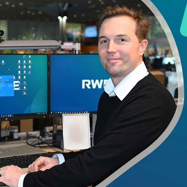 Ein Mann sitzt an einem Schreibtisch mit zwei Monitoren, auf denen das RWE-Logo und Desktop-Inhalte sichtbar sind.