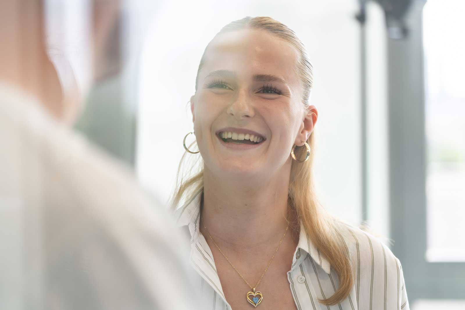 Eine Frau mit langen blonden Haaren trägt ein gestreiftes Hemd und eine herzförmige Halskette und schaut in einen Spiegel.