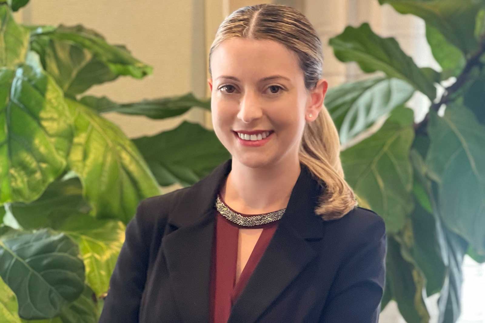 Eine Person in einem eleganten Schwarz mit einem geschmückten Kragen steht vor großen, grünen Blättern.