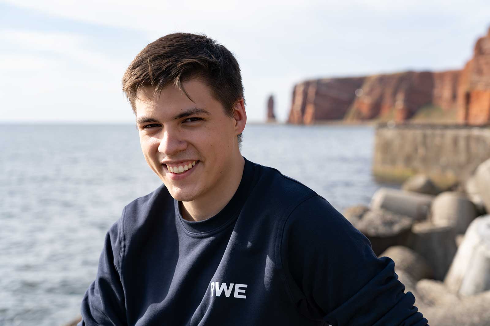 Eine Person sitzt am Wasser, trägt einen dunkelblauen Pullover mit der Aufschrift 'RWE' und Klippen im Hintergrund.