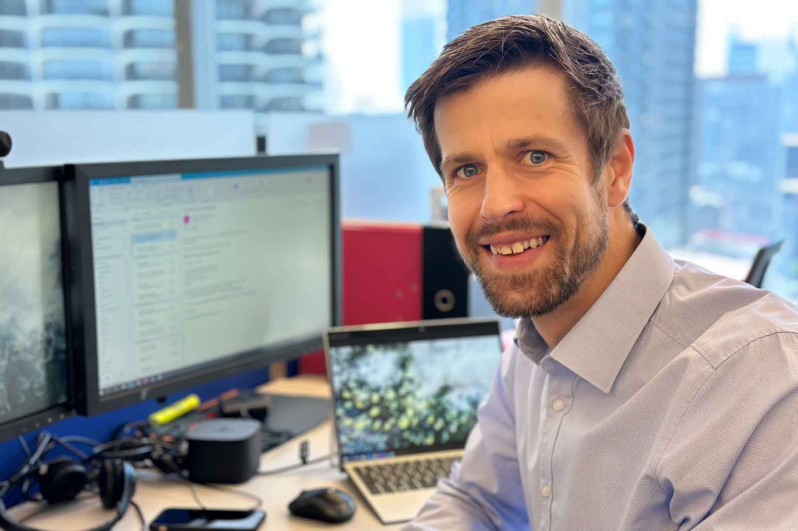 A man sits in an office with multiple monitors, working on a laptop. The cityscape is visible in the background.