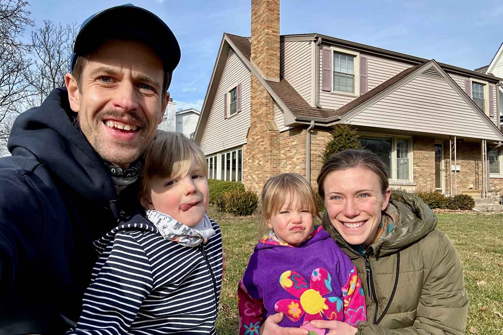 A family poses in front of a house, two adults holding children, grass in the foreground, and trees in the background.