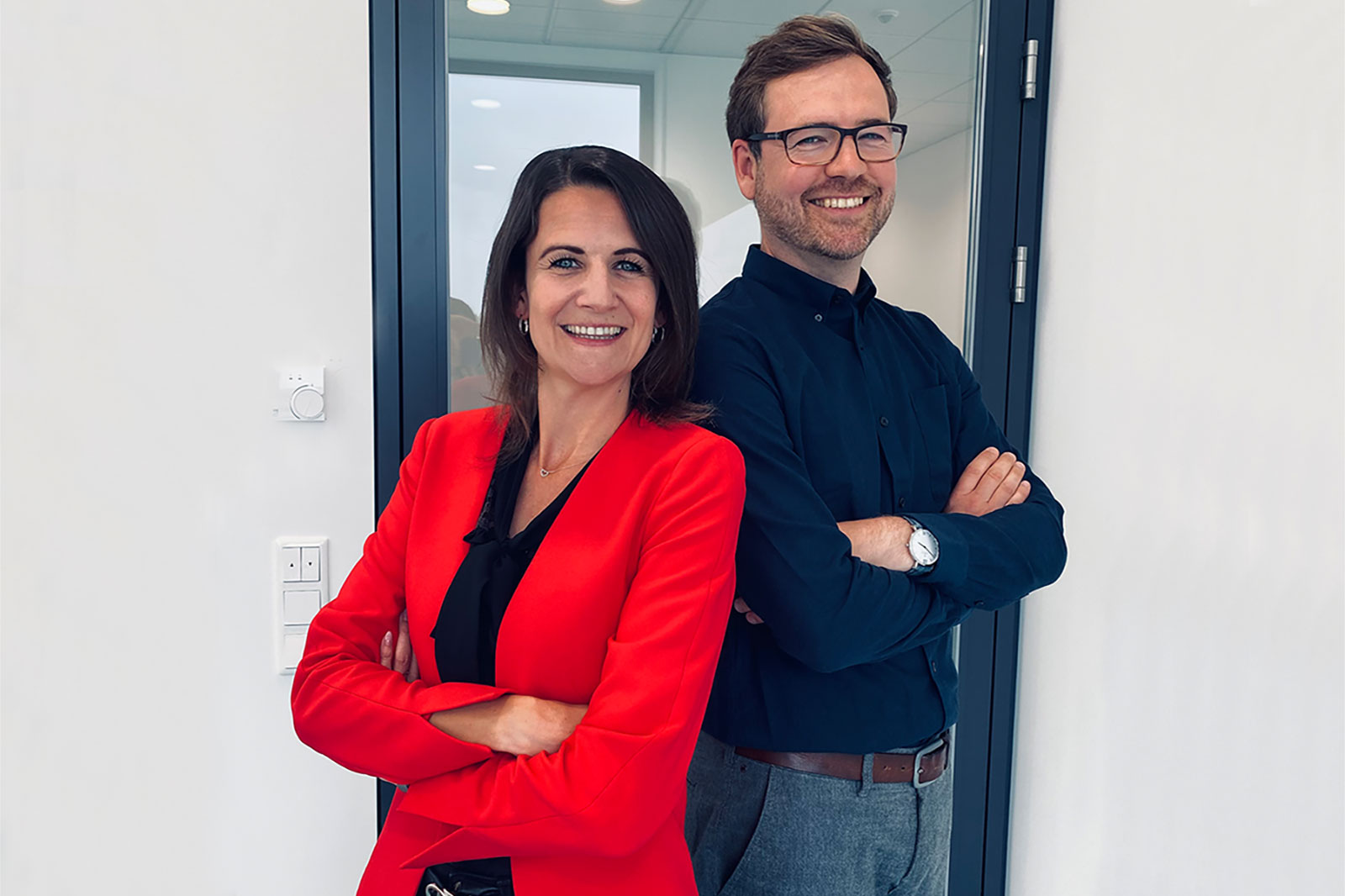 A man and a woman stand with arms crossed in a modern office in front of a glass panel.