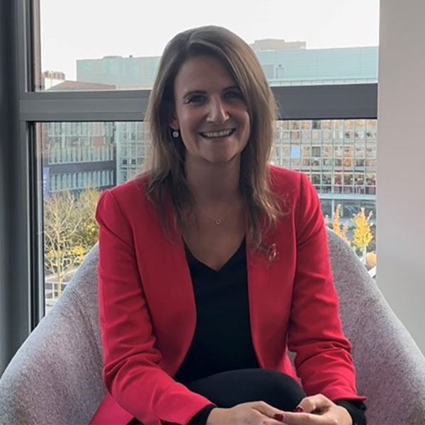 A person sits in a modern room with large windows. They are wearing a red blazer and have brown hair.
