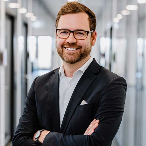 Ein Mann in einem Anzug steht mit verschränkten Armen in einem modernen Büro mit Glaswänden.