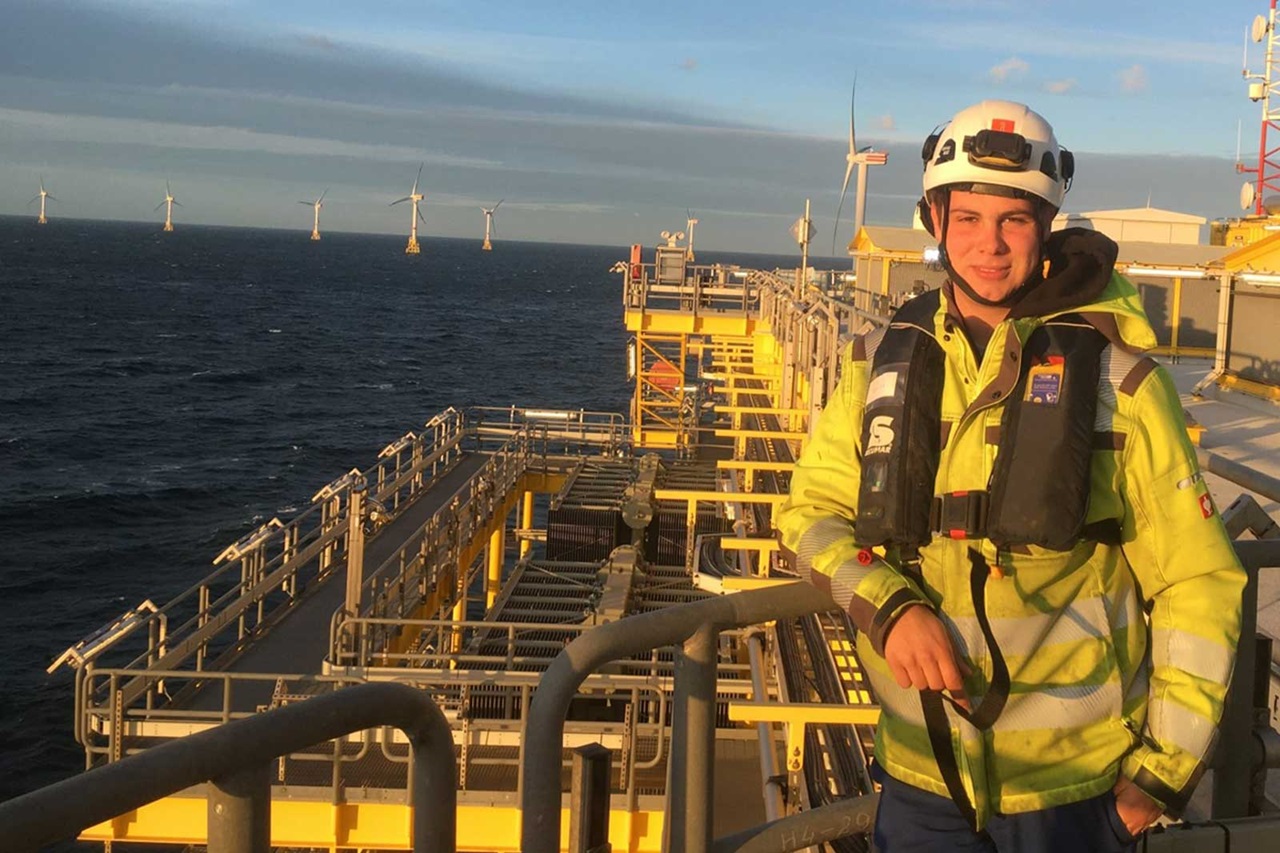 A worker stands on a platform overlooking the sea, with wind turbines visible in the background.