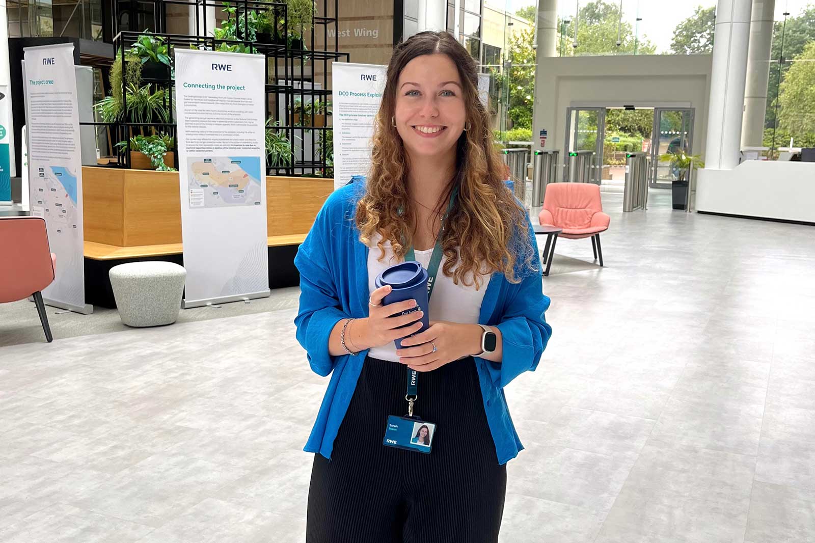 Eine Person steht in einem modernen Raum mit Pflanzen und Informationsständen. Sie hält einen Becher und trägt eine blaue Jacke.