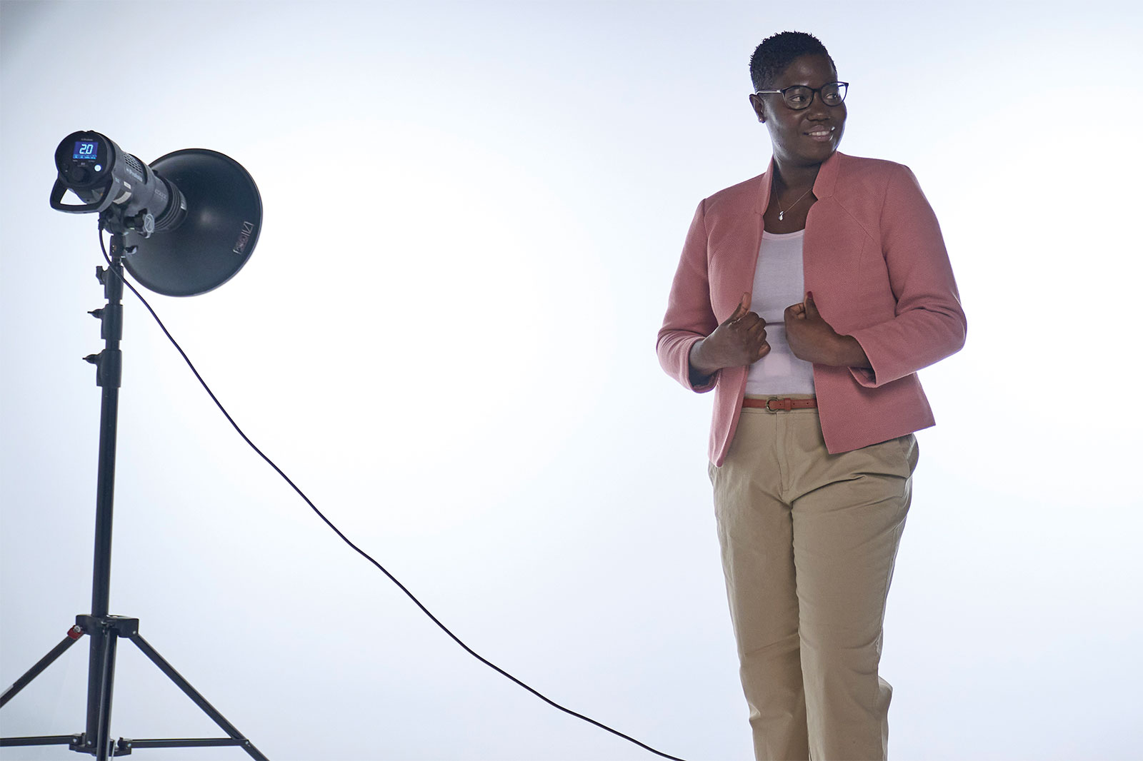 A person stands in front of a bright background. They are wearing a pink blazer and beige trousers.