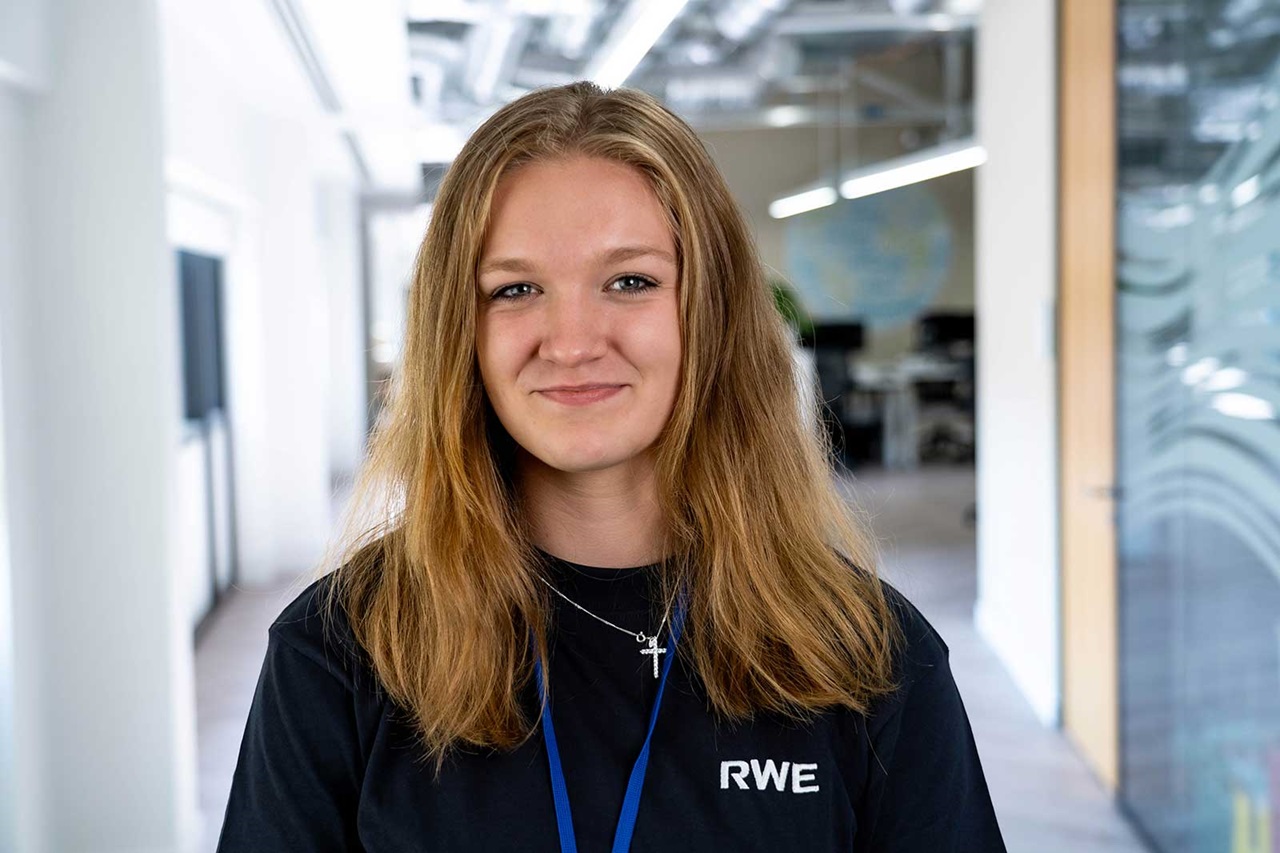 A person with long hair wearing a black RWE t-shirt stands in a bright, modern office environment.