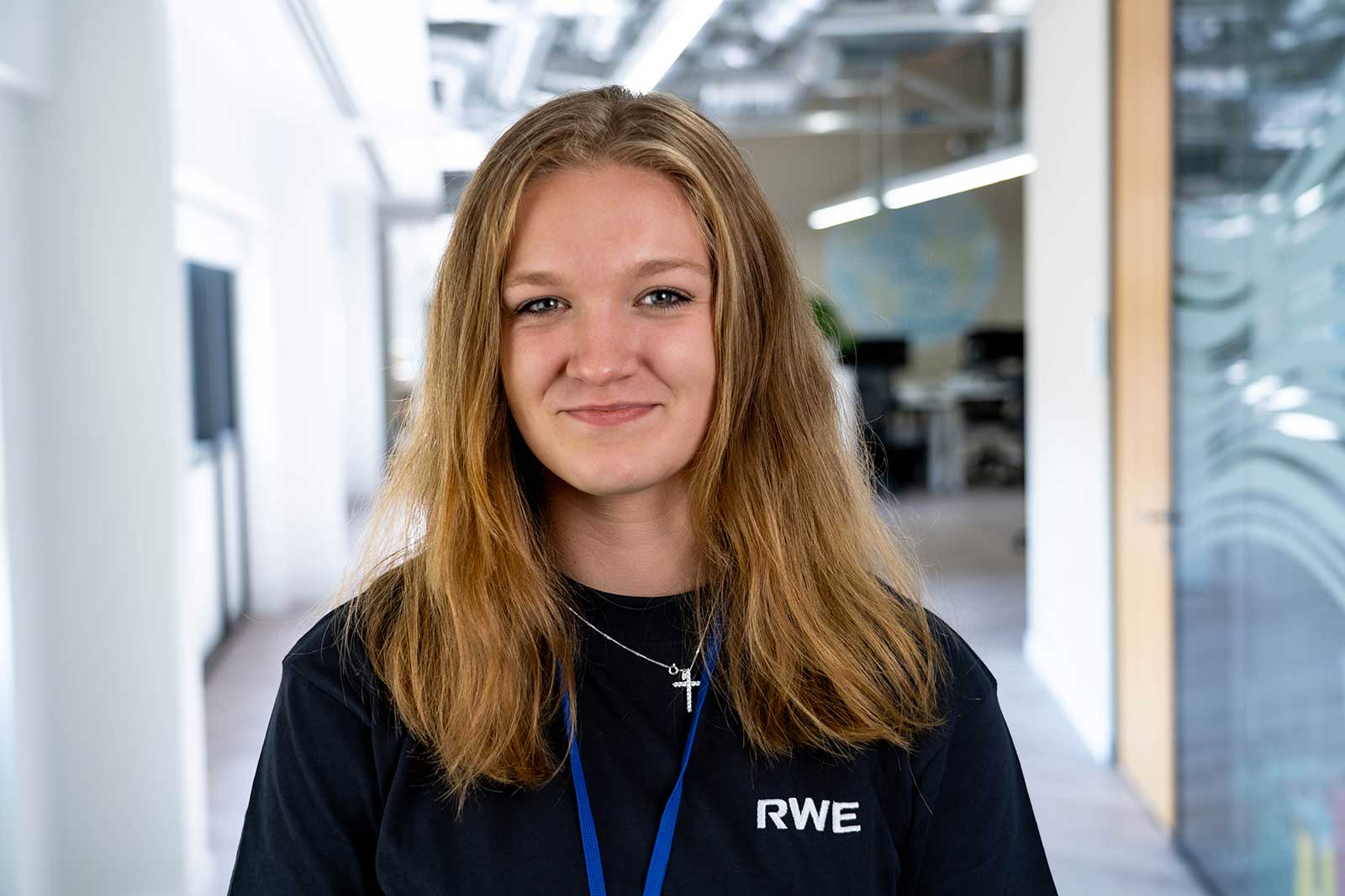A person with long hair wearing a black RWE t-shirt stands in a bright, modern office environment.