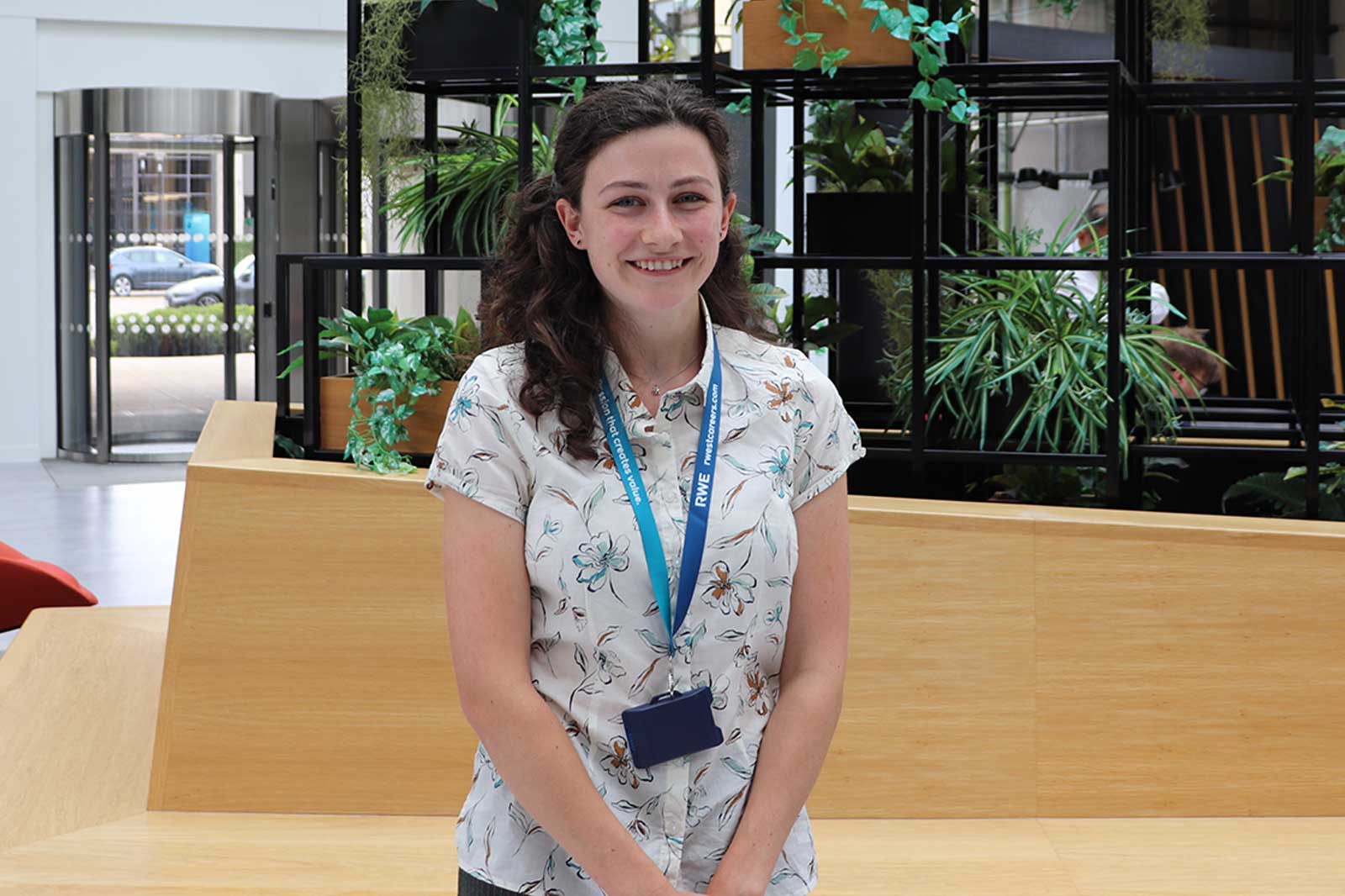 A woman wearing a floral shirt and ID badge stands in a modern indoor space surrounded by plants.