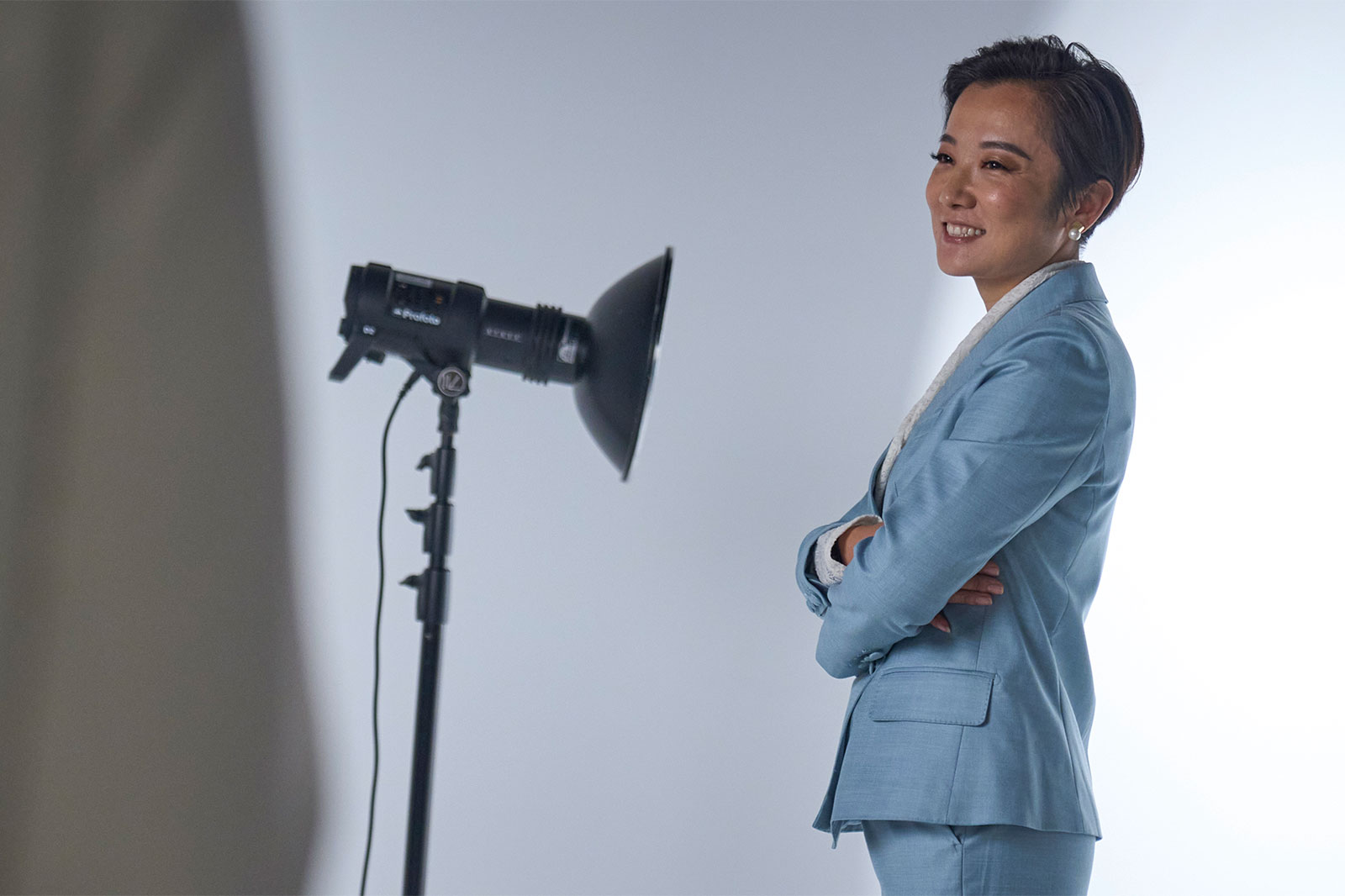 A person in a light blue suit stands with their arms crossed next to a professional lighting device.
