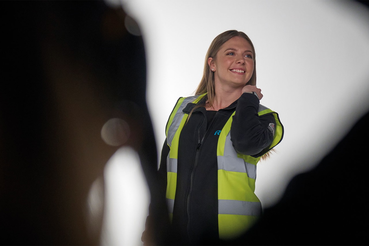 A person is wearing a reflective vest and stands in a studio environment with diffused lighting.