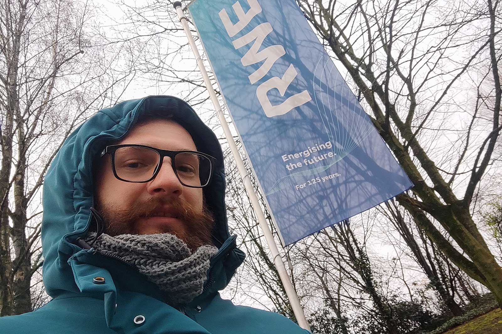A person is wearing a green jacket and a scarf, with an RWE flag fluttering in the background, surrounded by trees.