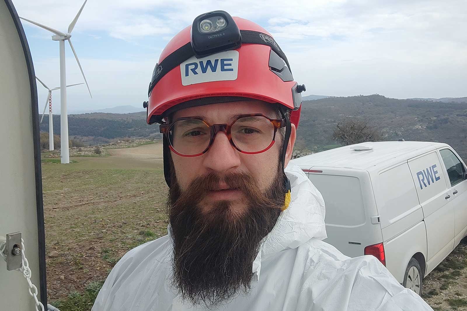 A technician in a white protective suit wearing a red helmet, with wind turbines and an RWE vehicle in the background.
